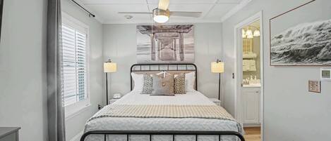 A bedroom with a bed, bedside lamps, a ceiling fan, a window with blinds, and a door leading to a bathroom.