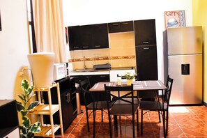A compact kitchen with black cabinets, a stainless steel refrigerator, a dining table with chairs, and a potted plant. - Lolos Place (Antigua Guatemala)