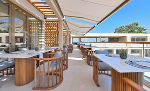 An outdoor dining area with wooden furniture and tables, large windows, and a view of the sea. - Pearl Island Chios Hotel Spa (Chios)
