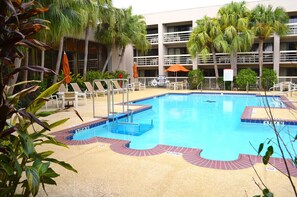 Una piscina con sillas de descanso, palmeras y un edificio de varios pisos al fondo.