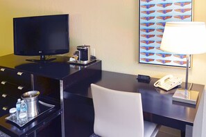 Habitación de hotel con escritorio, televisor, cafetera, teléfono, lámpara y una obra de arte en la pared.