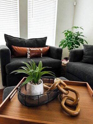 A living room with a sofa, a potted plant, a tray with a wooden sculpture, and a window with blinds. - Welcome to The Stagecoach Home – Your Texas Home Away From Home! (Rhome)