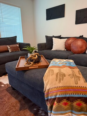 Un soggiorno moderno con un divano grigio, un vassoio di legno con una pianta, una coperta decorata e un tappeto marrone.