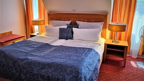 Quarto de hotel com cama, duas mesas de cabeceira, painel de cabeceira em madeira e cortinas.