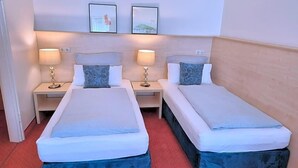 Quarto de hotel com duas camas, cada uma com cabeceira, mesas de cabeceira e luminárias. Há quadros nas paredes acima das camas.