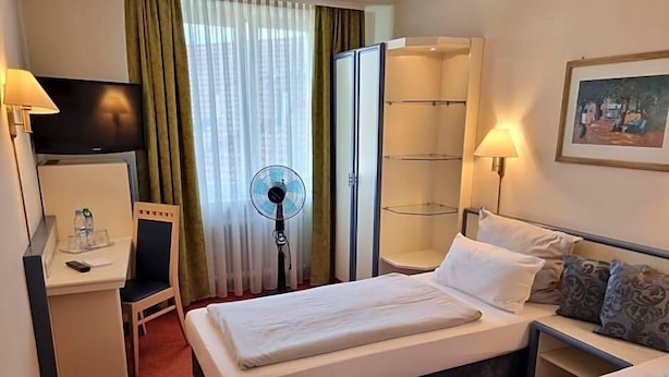 Quarto de hotel com duas camas, uma mesa com cadeira, televisão, ventilador, guarda-roupa e um quadro na parede.