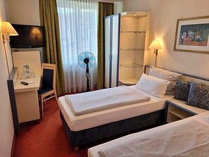 Une chambre d’hôtel avec deux lits, un bureau avec une chaise, une télévision, un ventilateur, une armoire et un tableau au mur.