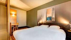 A hotel room with a large bed, a desk with a chair, a window with a view of a cityscape, and a wall-mounted light. - Hotel Premia Re Classe Roissy Saint Witz (Saint-Witz)