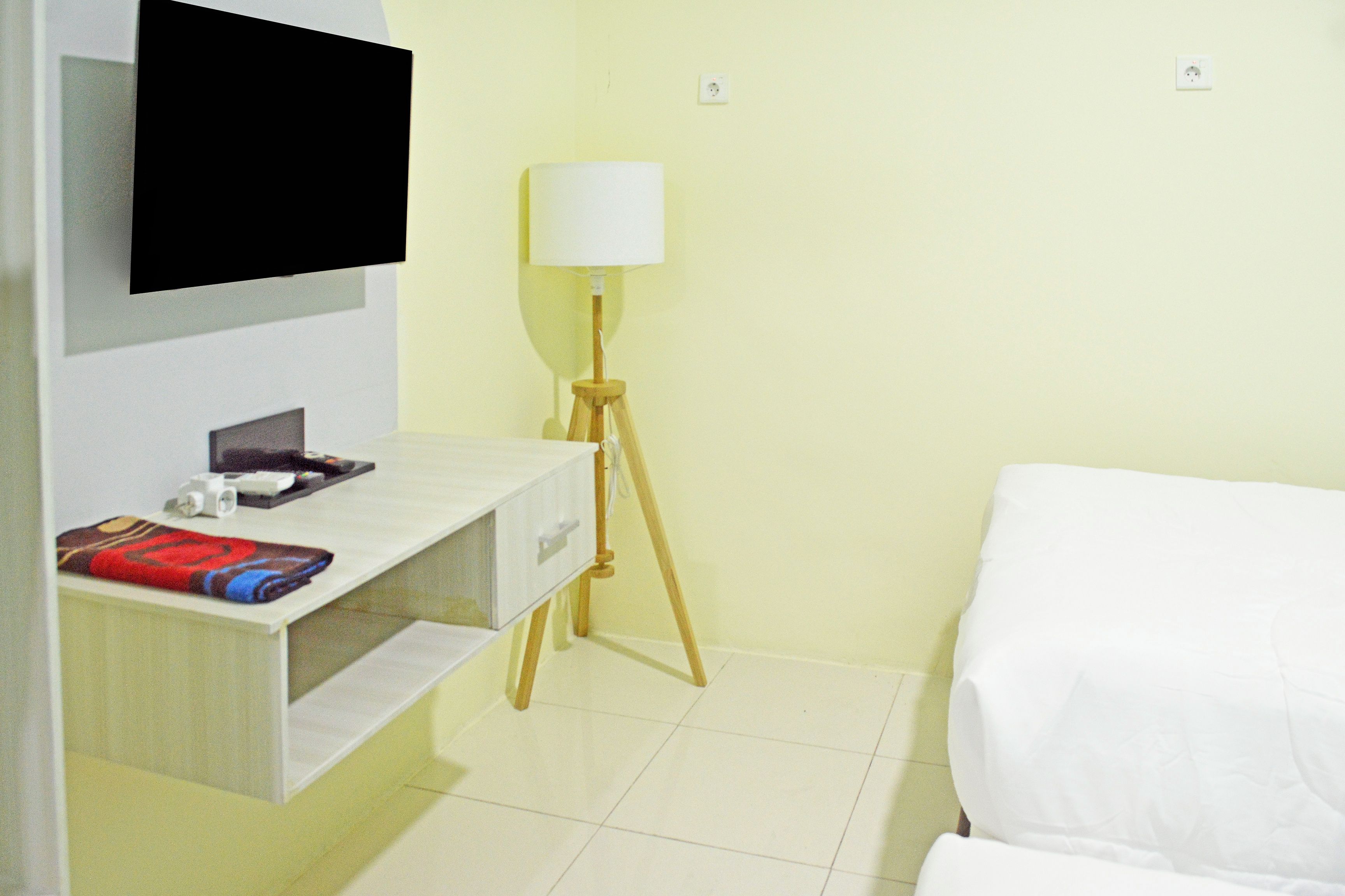 Habitación de hotel con una cama, televisor montado en la pared, mesita de noche con lámpara y una pequeña estantería con una manta.