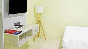 Habitación de hotel con una cama, televisor montado en la pared, mesita de noche con lámpara y una pequeña estantería con una manta.