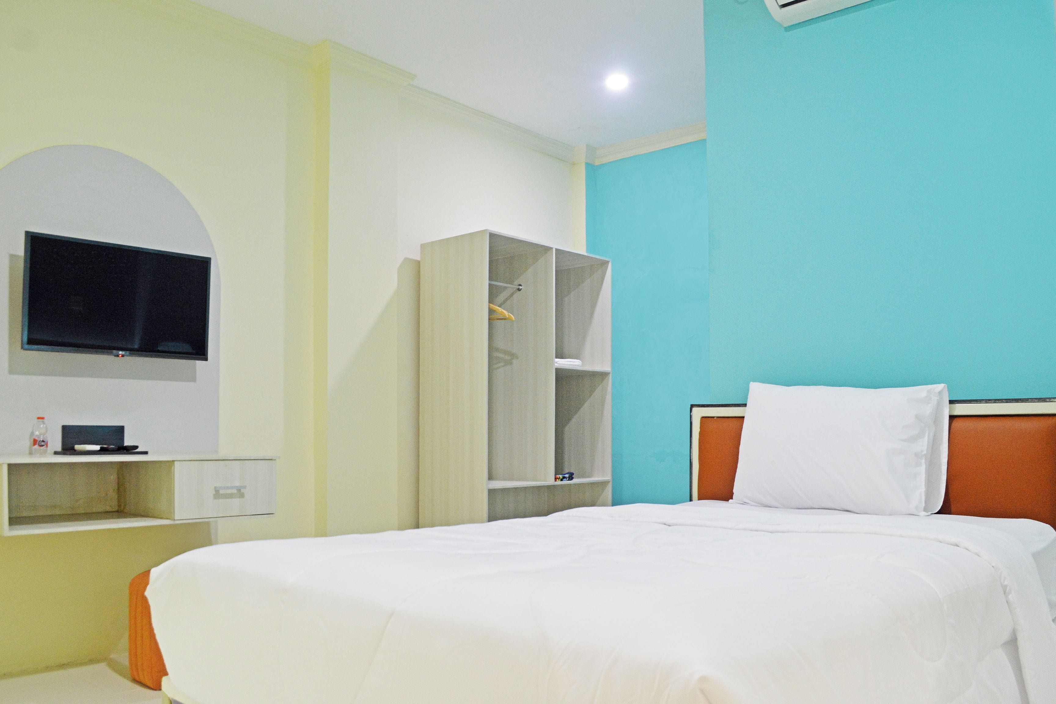 A hotel room with a bed, a television mounted on the wall, a wardrobe, and a shelf with a bottle.