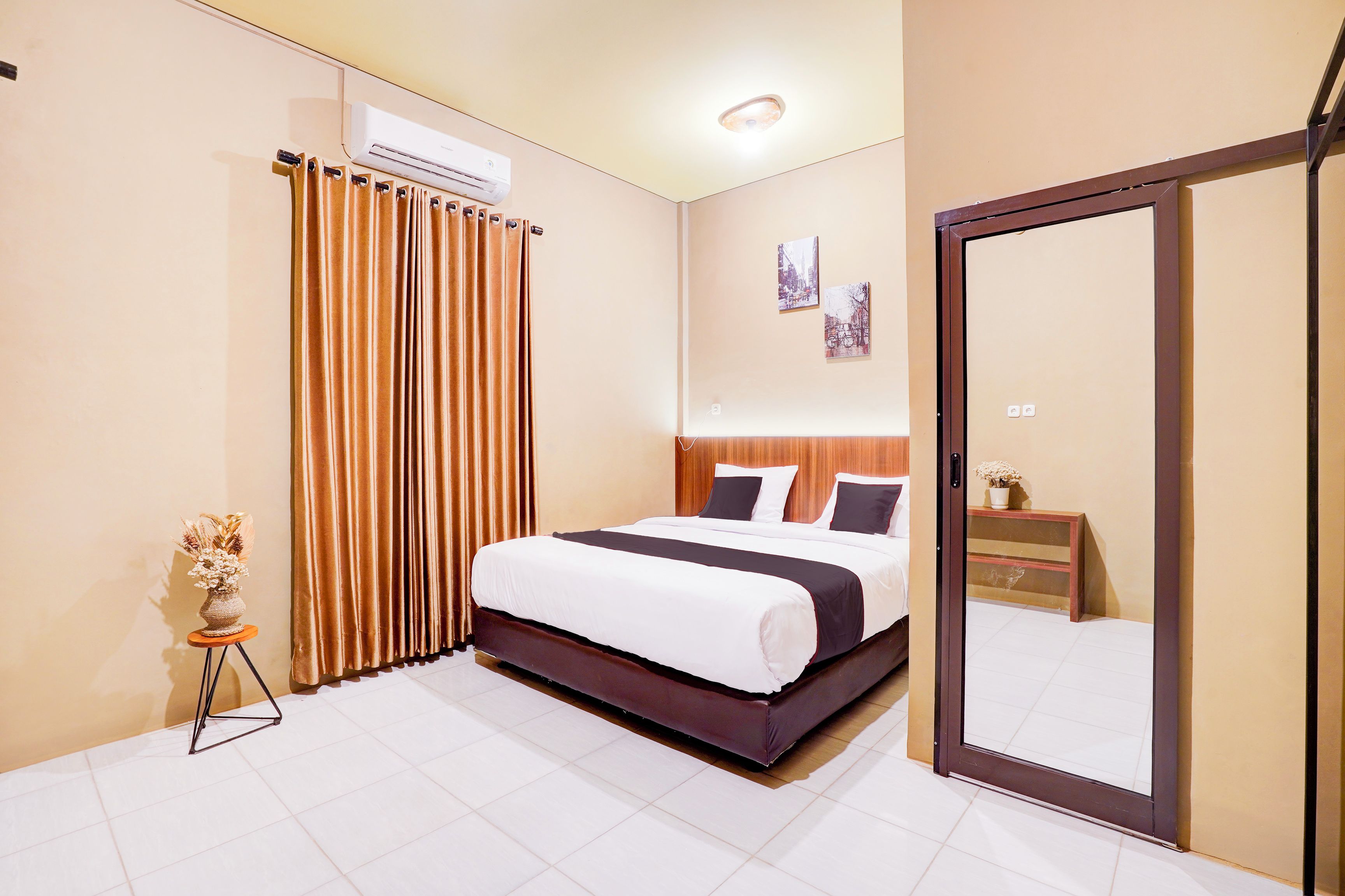 A hotel room with a bed, a mirror, a small table with a vase, and a window with curtains.