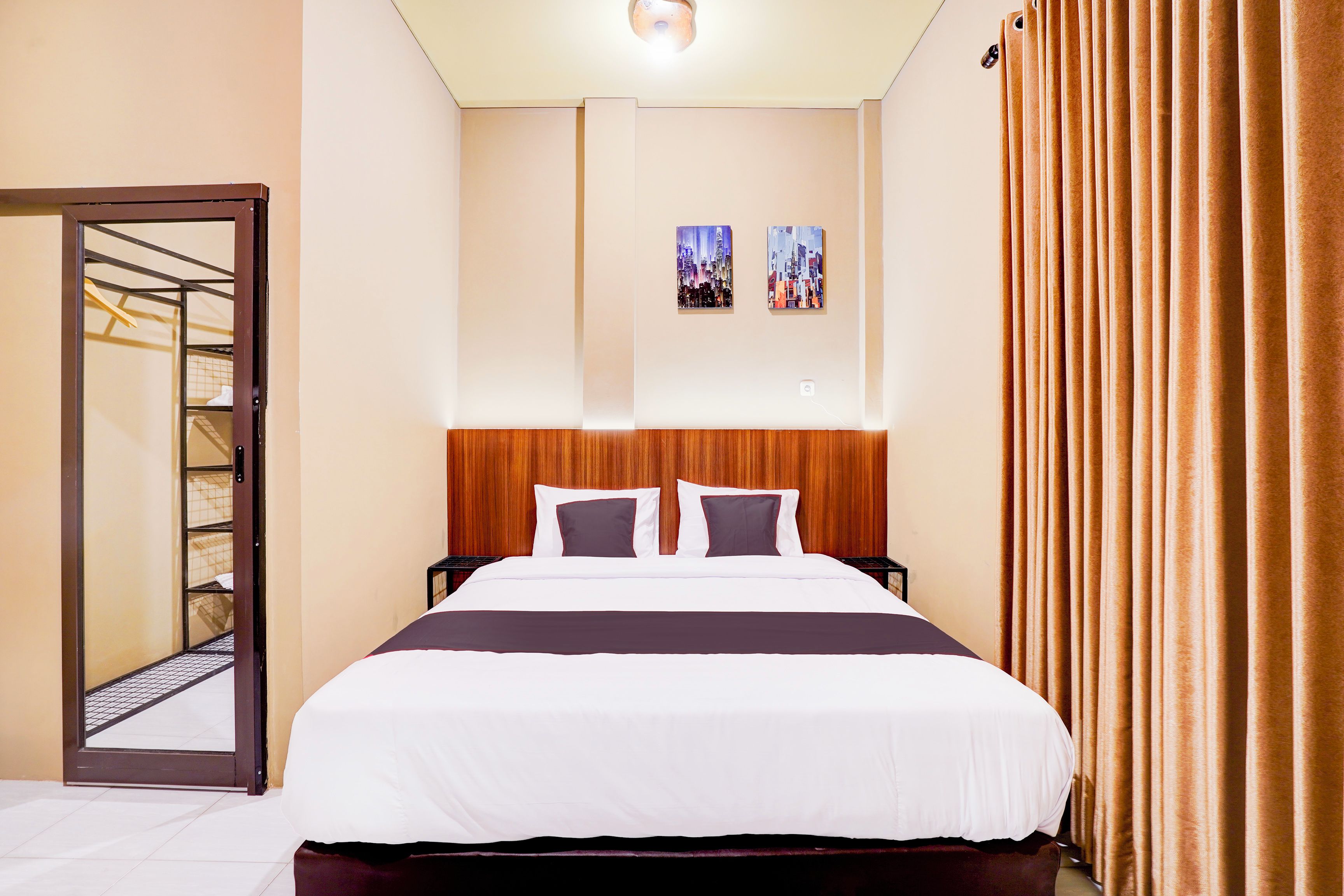 Habitación de hotel con una cama grande, cabecera de madera, dos cuadros en la pared y vista a un paisaje urbano por la ventana.
