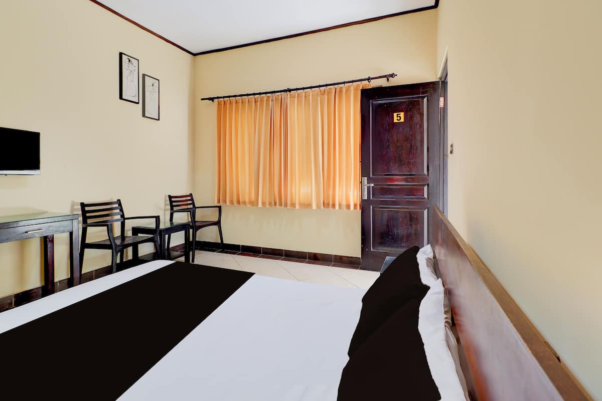 A hotel room with a bed, a desk, chairs, a window with curtains, and a door with a number.