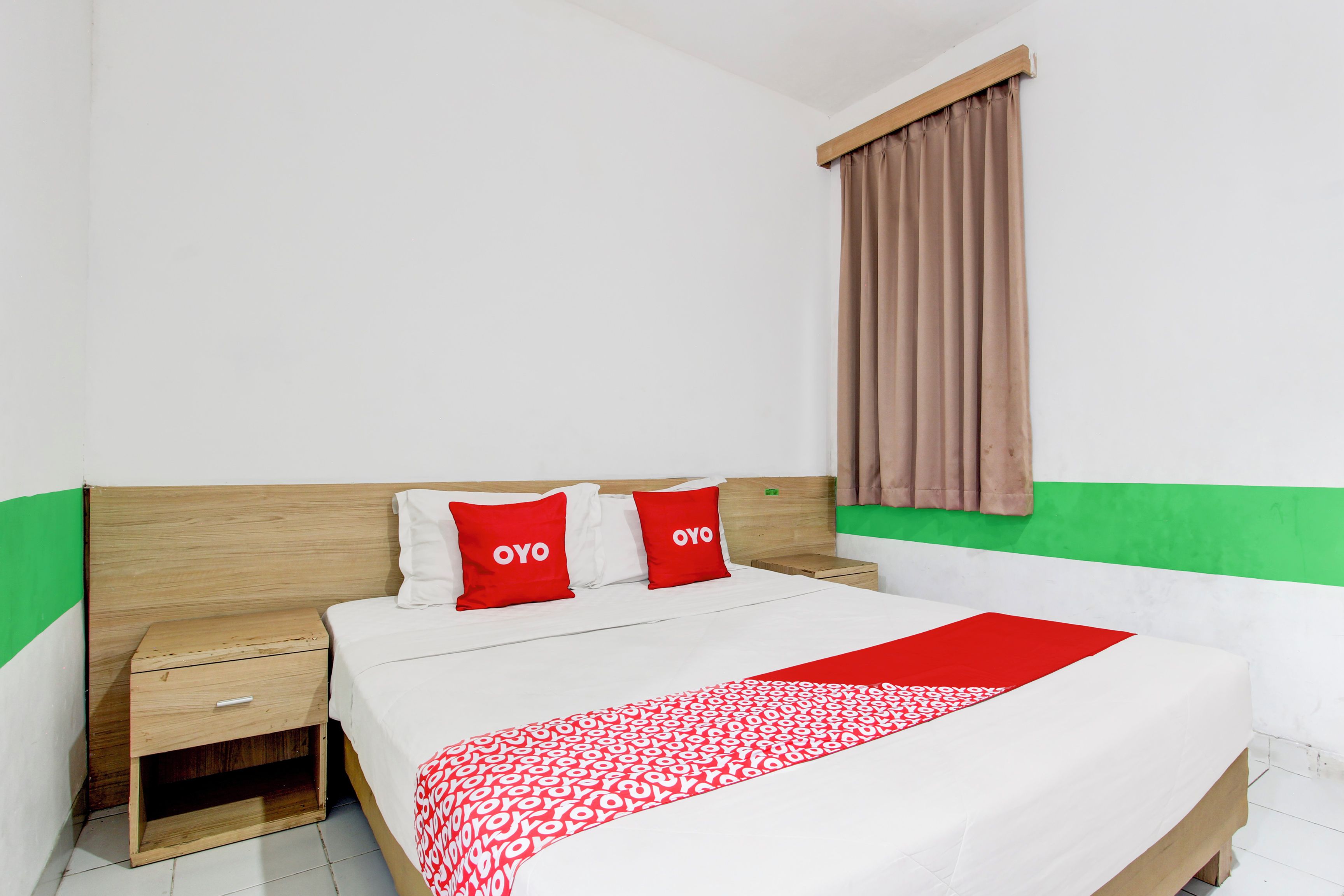A neatly made bed with a red and white patterned bedspread and two red pillows with "OYO" text. There is a wooden nightstand with a drawer and a small shelf below it. The room has white walls with a green stripe at the bottom and a window with brown curtains.