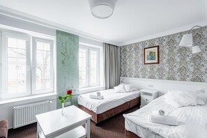 A hotel room with two beds, a small table, a radiator, and a window with curtains.