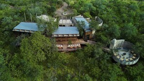 An aerial view of a secluded lodge surrounded by dense forest, featuring a central building with a sloped roof, a circular outdoor seating area, and a small circular structure with a dome roof. - Bupenyu Luxury Boutique Lodge (Matabeleland North)