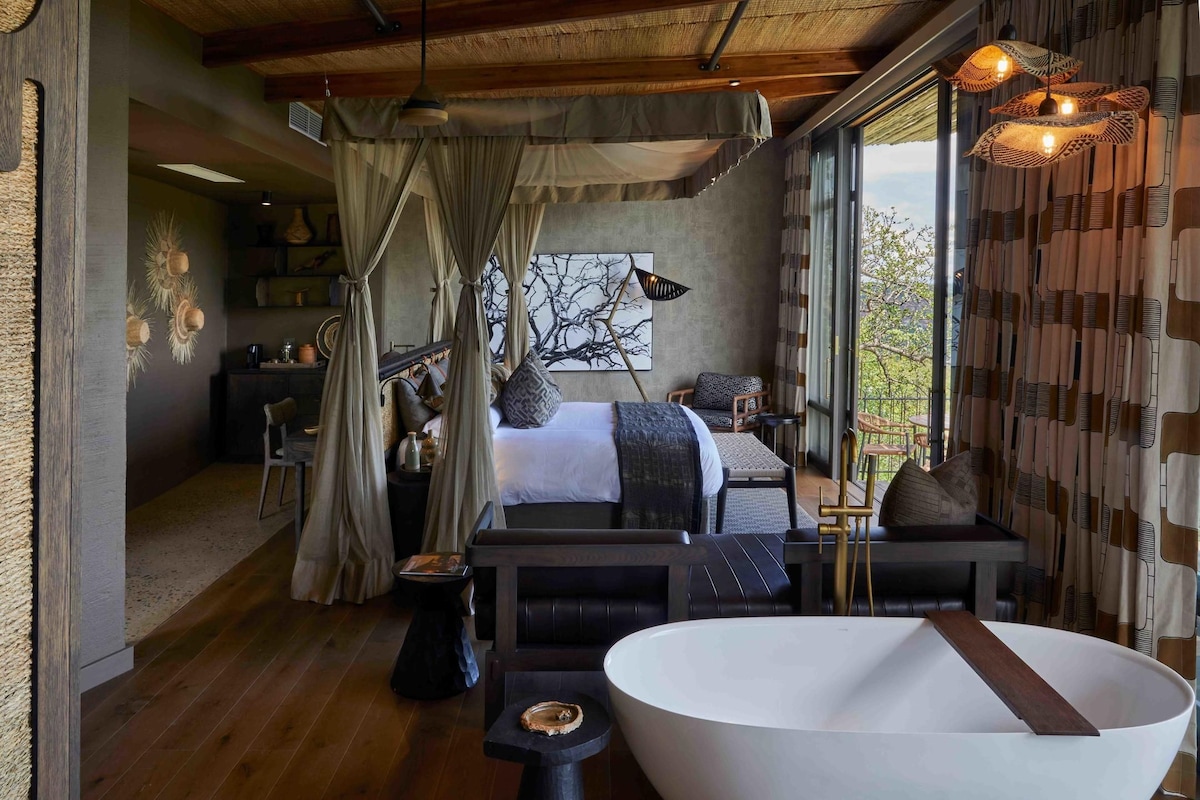 A modern bedroom with a large bed, a sitting area, a bathtub, and a view of the outdoors.