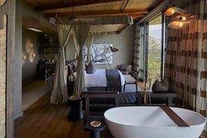 A modern bedroom with a large bed, a sitting area, a bathtub, and a view of the outdoors. - Bupenyu Luxury Boutique Lodge (Matabeleland North)