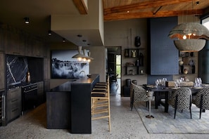 A modern kitchen with a central island, bar seating, and a dining area with patterned chairs. - Bupenyu Luxury Boutique Lodge (Matabeleland North)