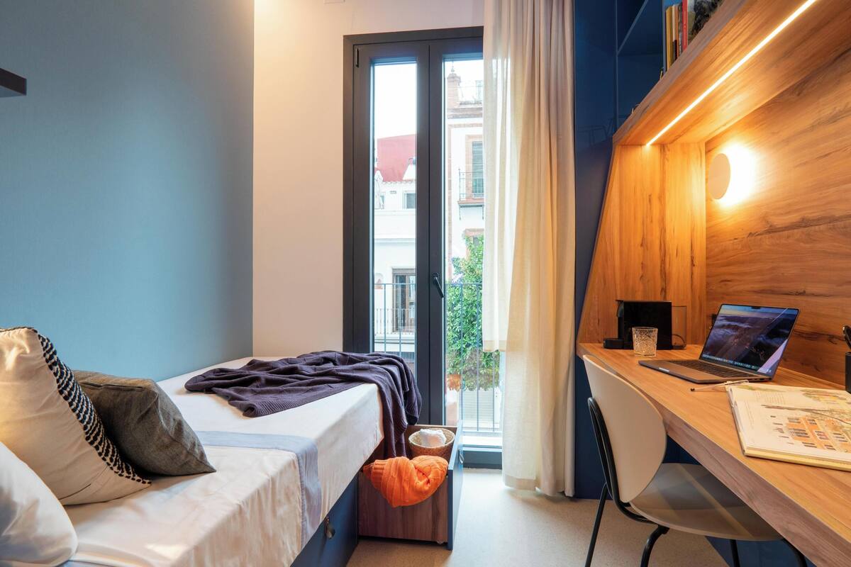 Quarto de hotel com uma cama, uma escrivaninha com um laptop, uma janela com cortinas e vista para edifícios.