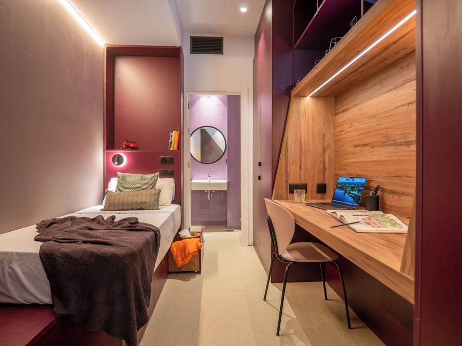 Un dormitorio moderno con una cama individual, un escritorio con una computadora portátil, una silla y un baño pequeño con un espejo y lavamanos.
