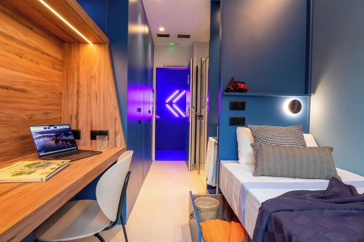 Quarto de hotel moderno com escrivaninha de madeira, um laptop, cama com almofadas e parede com detalhes em azul.