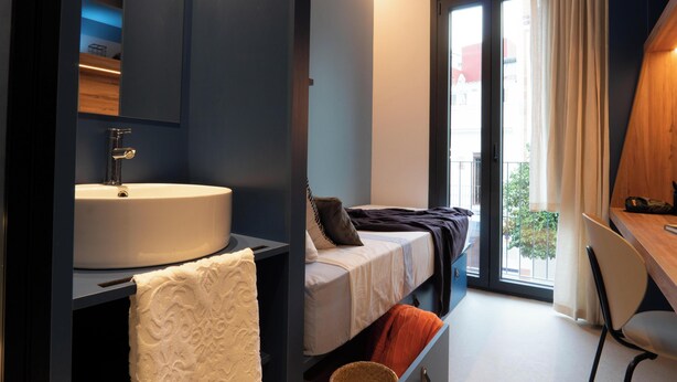 Quarto de hotel moderno com uma pia grande, uma cama com almofadas, uma cadeira e vista para o exterior.