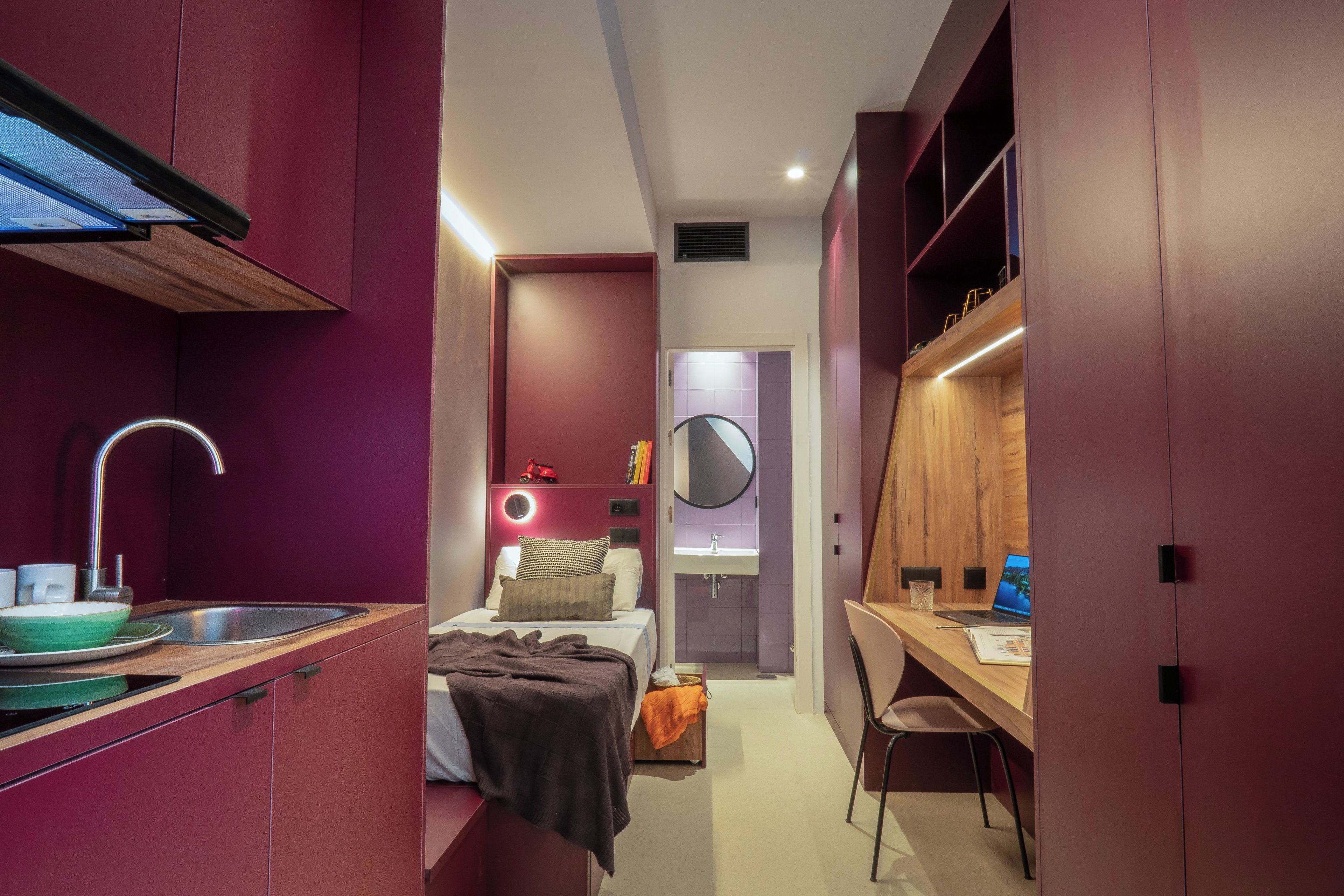 A compact bedroom with a bed, bedside table, and a desk with a chair. There is a built-in wardrobe and a small kitchenette with a sink and a bowl.