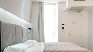 Una habitación de hotel con cama, puerta, ventana con cortinas y una lámpara fijada a la pared.