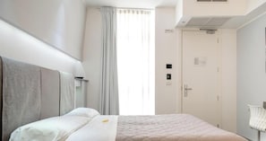 A hotel room with a bed, a door, a window with curtains, and a wall-mounted light fixture. - Hotiday Brindisi Vittoria  (Brindisi)