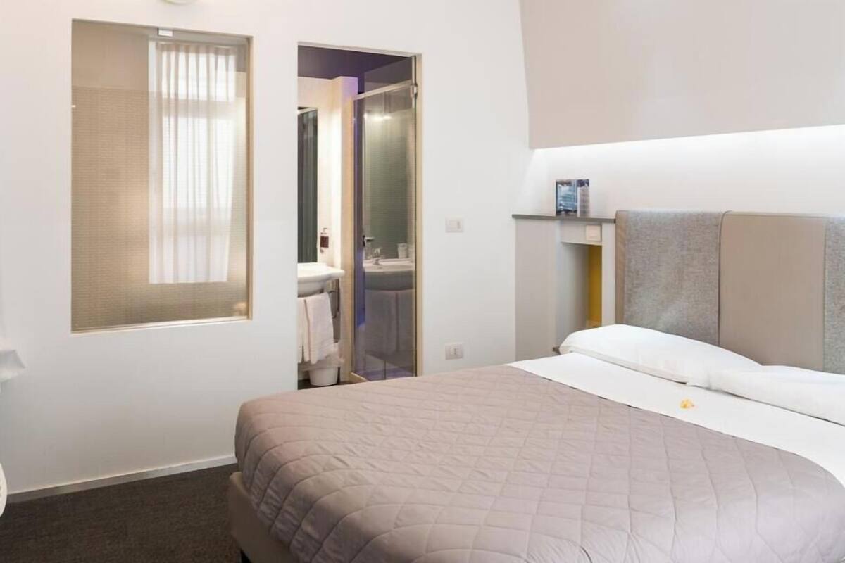 Habitación de hotel con cama, ventana con persianas, zona de ducha y un pequeño estante con artículos de aseo.