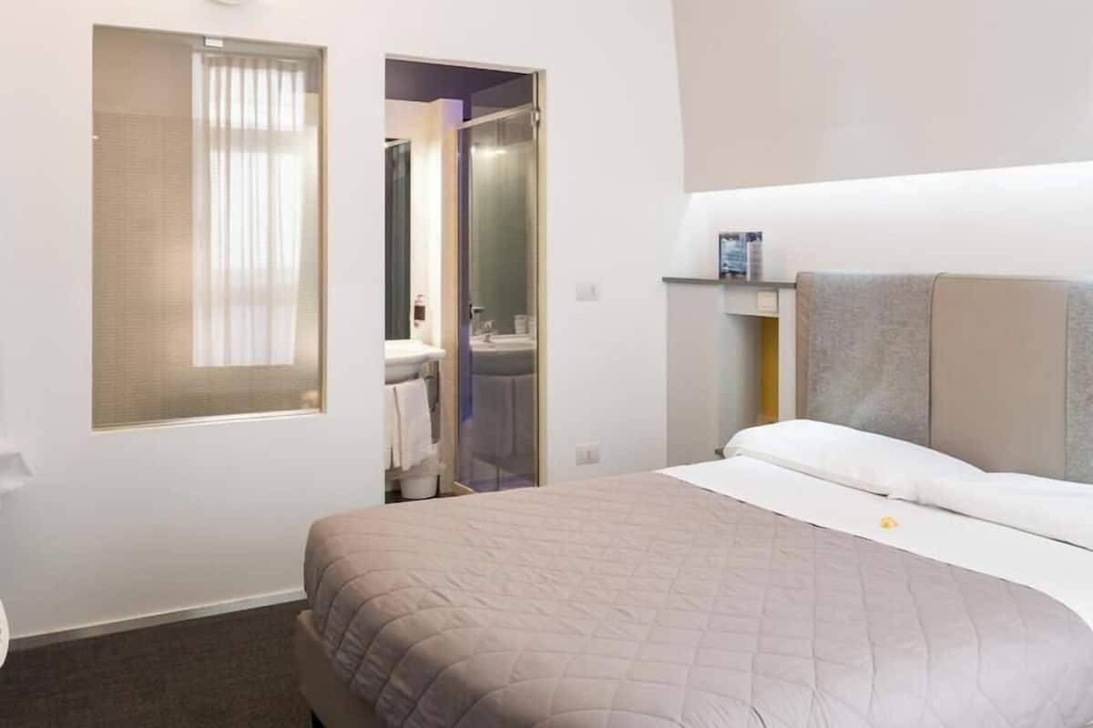 Habitación de hotel con cama, ventana con persianas, zona de ducha y un pequeño estante con artículos de aseo.