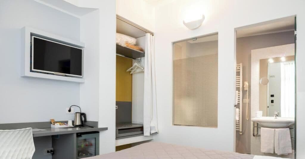 Habitación de hotel con cama, escritorio con hervidor, televisión, armario y baño con espejo y toallero.