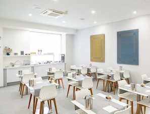 A modern conference room with white tables and chairs, a kitchen area, and two large abstract wall art pieces.