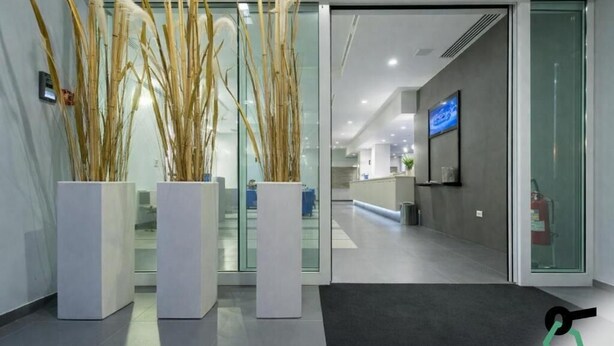 Zona de recepción con puerta de cristal, plantas en macetas, mostrador de recepción y extintor.