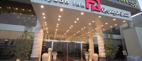 The exterior of Riyadh Inn with a prominent R logo, a covered entrance, and a glass facade.