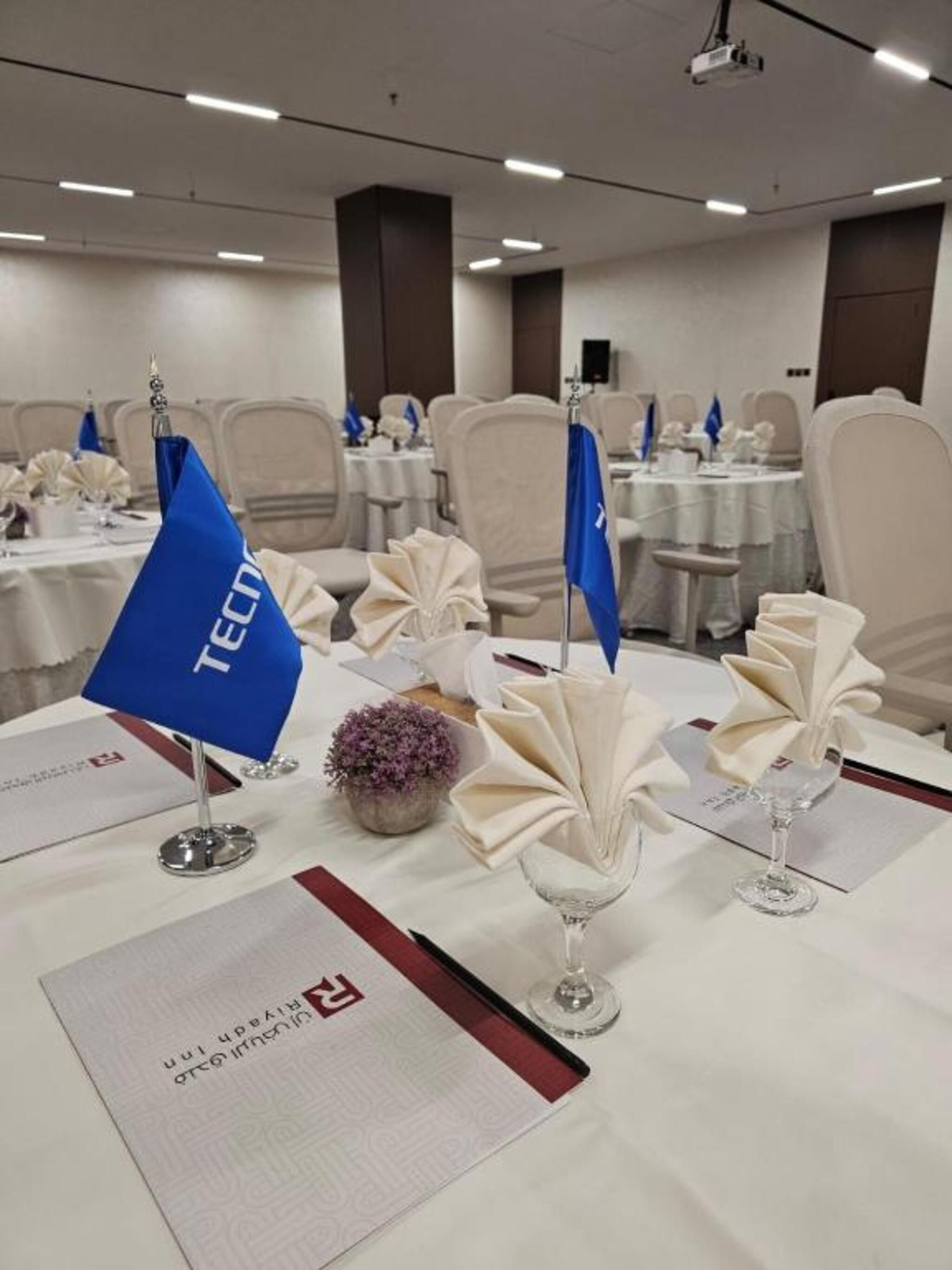 Un salón de conferencias con mesas dispuestas para una reunión, con tarjetas de identificación, manteles y un proyector.