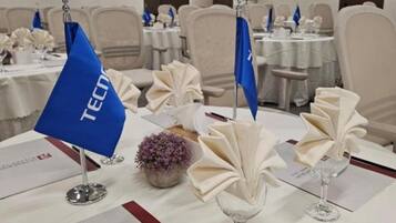 Un salón de conferencias con mesas dispuestas para una reunión, con tarjetas de identificación, manteles y un proyector.