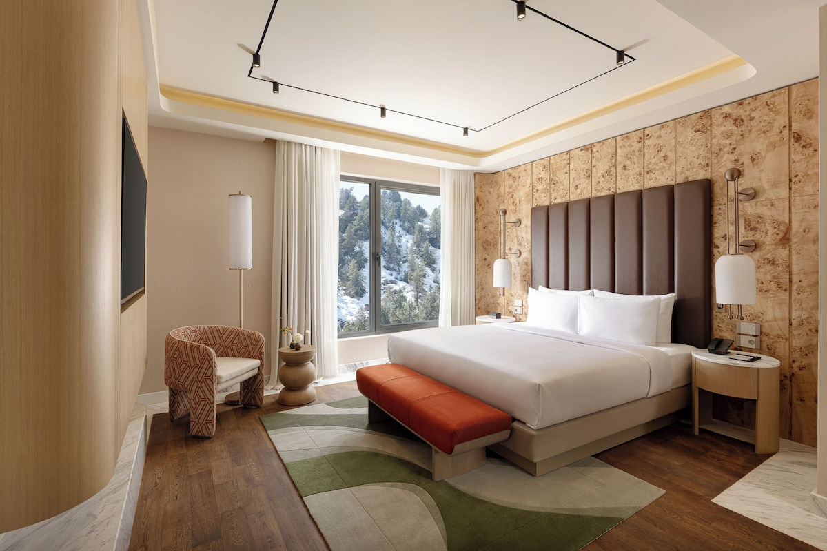 A modern hotel room with a large bed, a bench, a chair, a bedside table, and a view of a snowy landscape.