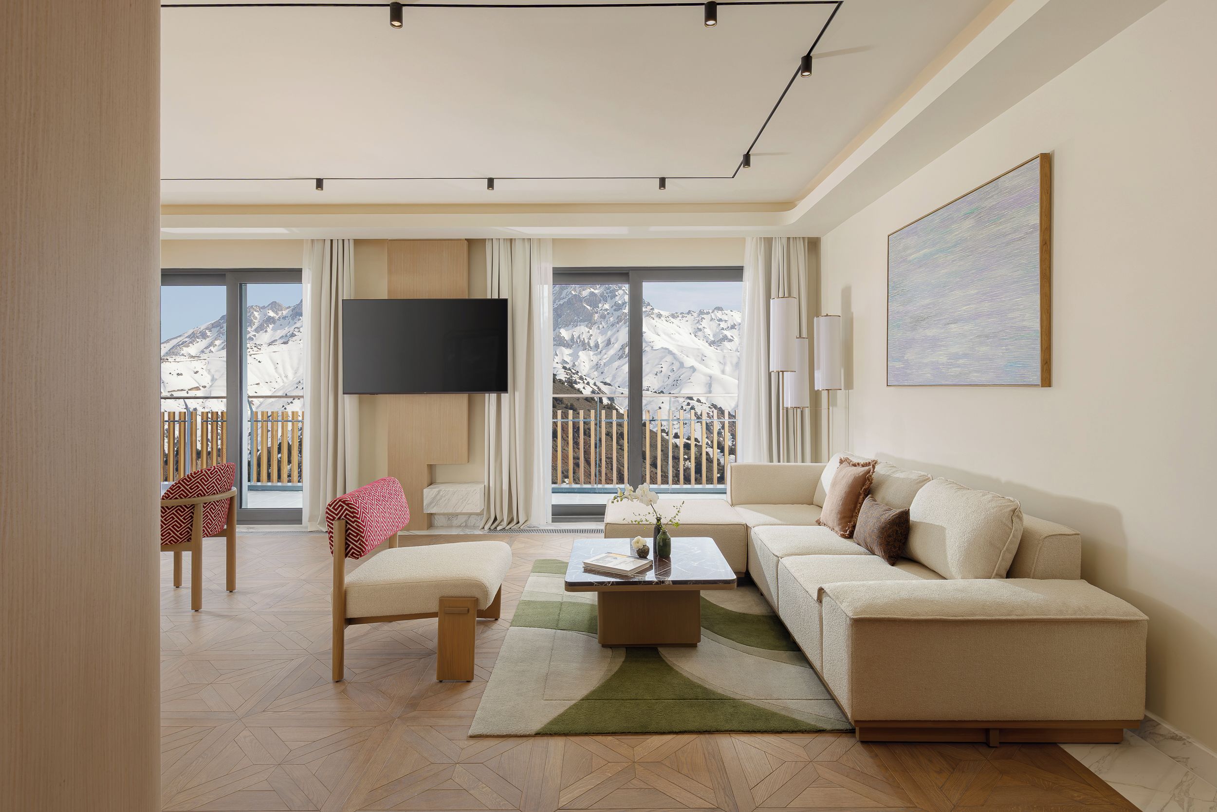 A modern living room with a sofa, a coffee table, and a TV. There are large windows with a mountain view.