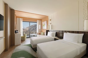 A hotel room with two beds, a dining area, a view of mountains, and a large window.