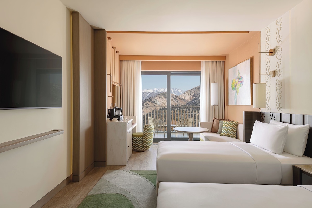 A hotel room with a bed, a desk, a chair, a table, and a view of mountains.