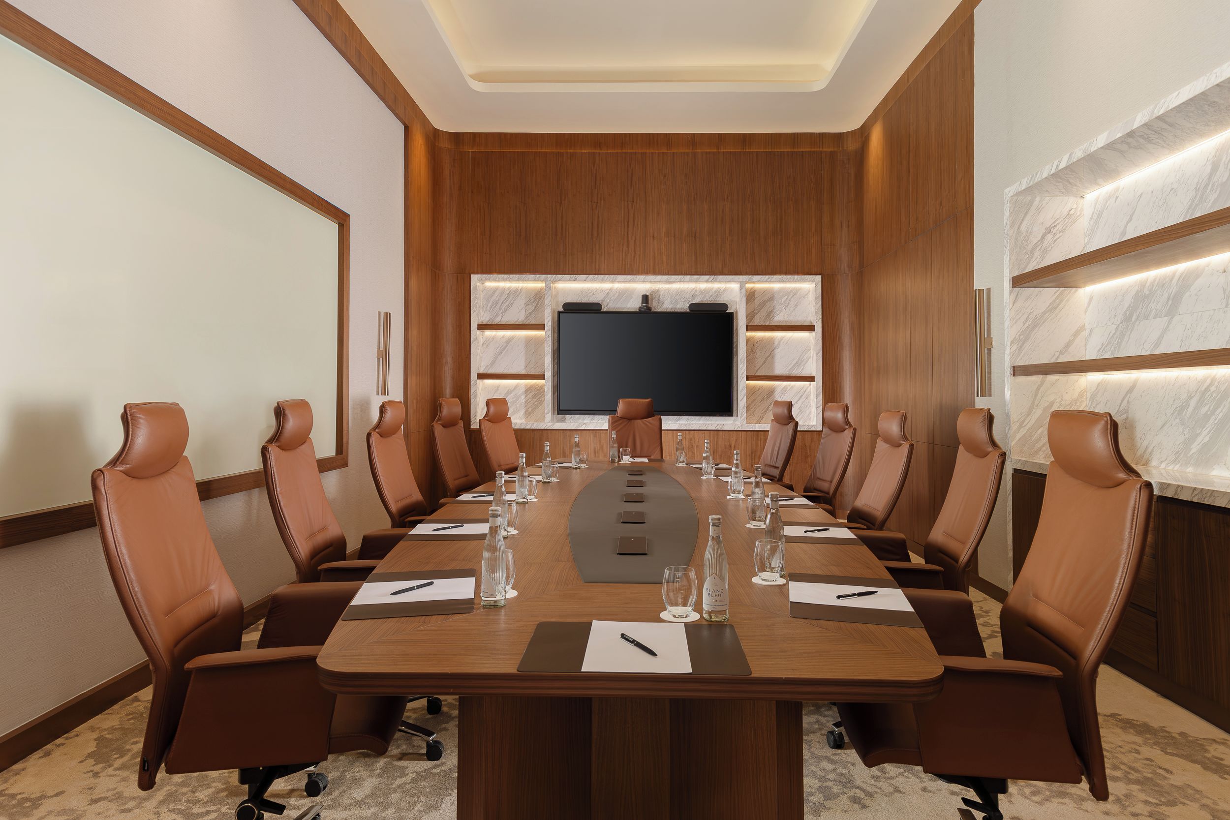 A conference room with a long table, brown leather chairs, a large flat-screen TV, and a marble accent wall.