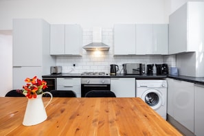 Eine moderne Küche mit weißen Schränken, einem Holztisch, einer weißen Vase mit Blumen, einem Gasherd, einer Mikrowelle, einer Kaffeemaschine und einer Waschmaschine.