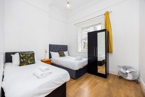 A hotel room with two beds, a mirror, a fan, and a window with a yellow curtain. - 5 mins from the Station.4Bedrooms Spacious Apartment  with Parking and Garden (London)