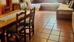 A rustic room with wooden walls, a tiled floor, a wooden dining table with chairs, a sofa, a bed, and a TV.