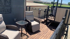 Una terraza techada al aire libre con mobiliario de mimbre, baranda metálica y vista a un campo verde y colinas lejanas.