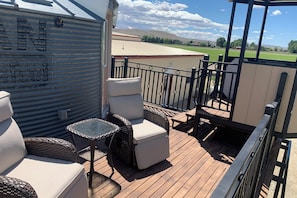Une terrasse extérieure couverte, aménagée avec des meubles en osier, une rambarde en métal et offrant une vue sur un champ verdoyant et des collines lointaines.