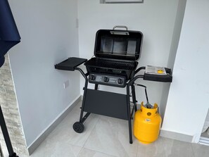 Ein tragbarer Grill mit Rädern, einer Gasflasche und einem kleinen Ablagefach mit einer Broschüre.